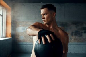 A shirtless man is performing a kettlebell exercise in sports medicine setting.